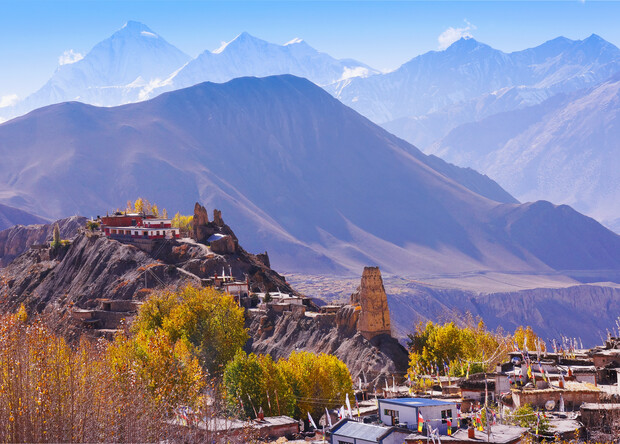 A monastery perches on a rocky hill, surrounded by autumnal trees and traditional houses, with towering snow-capped mountains in the distant background under a clear blue sky.