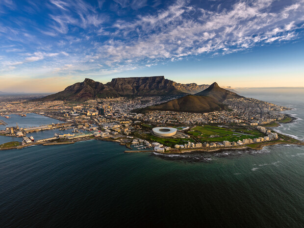 Aerial view of a coastal city with a prominent stadium, surrounded by buildings and mountains under a partly cloudy sky. The ocean borders the city, adding scenic beauty.