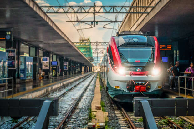 A sleek, modern train sits on railway tracks at a busy station, flanked by platforms with people and signage. Overhead wires and a cloudy sky are visible.