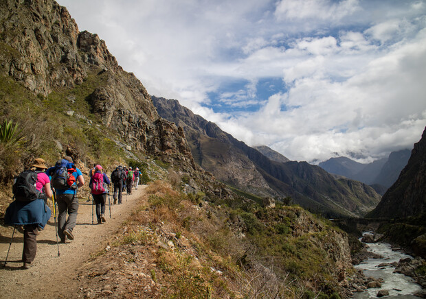 Hikers walk along a rugged mountain trail, using trekking poles. They are surrounded by steep, rocky cliffs under a partly cloudy sky, with a river flowing in the valley below.