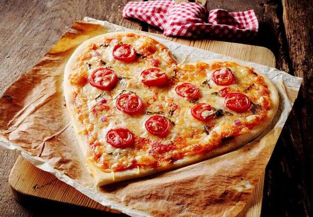 Heart-shaped pizza topped with cherry tomatoes and cheese rests on parchment paper, atop a wooden cutting board. A checkered cloth lies nearby on a rustic wooden table.