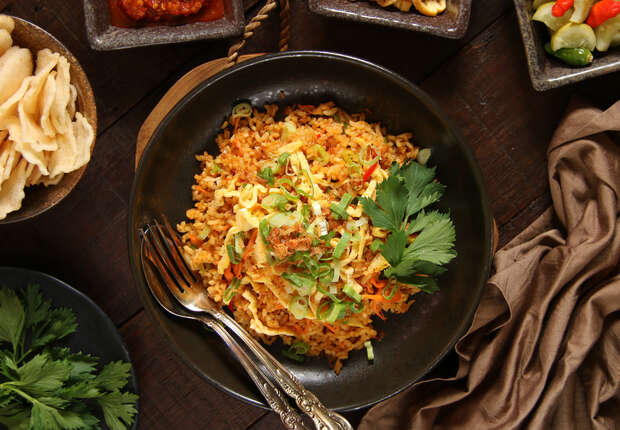 A bowl of fried rice topped with sliced vegetables and herbs sits on a wooden surface, surrounded by side dishes, condiments, and utensils.