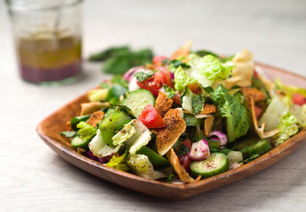 A wooden plate holds a colorful fattoush salad with lettuce, cucumbers, tomatoes, mint, and crispy flatbread pieces. In the background, a glass jar contains dressing.