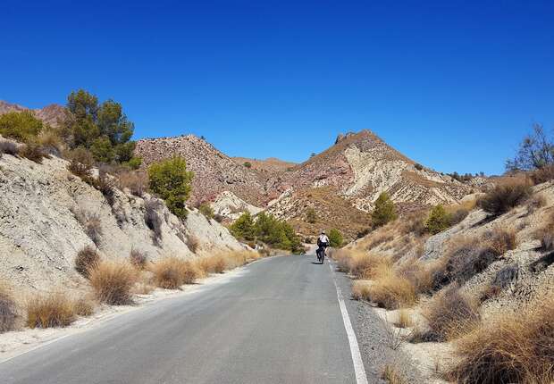 A cyclist rides along a paved road through a dry, mountainous landscape with sparse vegetation and clear blue skies overhead.