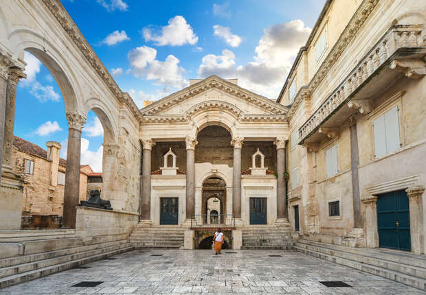 Ancient stone courtyard surrounds a seated archway structure with grand columns. A lone person walks in the center under a bright blue sky filled with fluffy clouds.