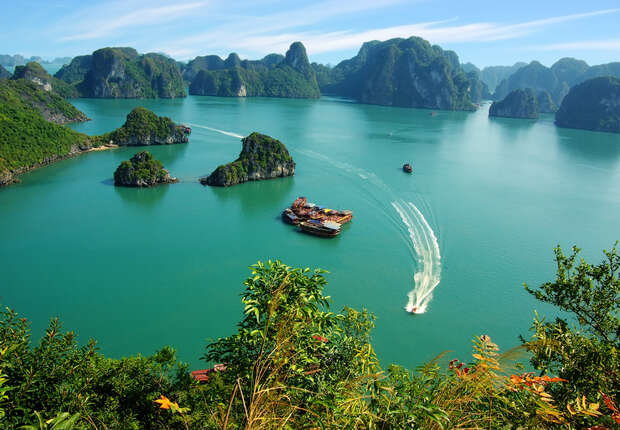 A small boat creates a white wake while navigating between lush, rocky islets in a serene, expansive turquoise bay, surrounded by dense greenery under a clear blue sky.