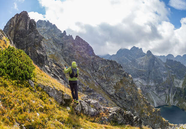 A hiker in a green jacket stands on a rocky path, overlooking a mountainous landscape with steep cliffs and a distant lake under a partly cloudy sky.