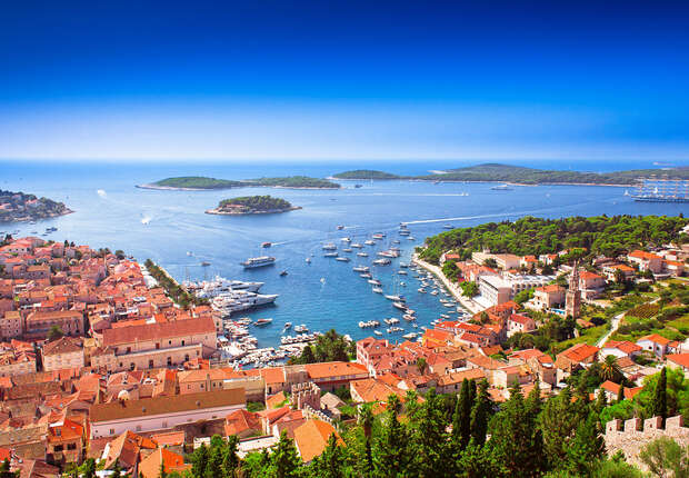 Boats float in a vibrant blue bay, surrounded by a coastal town with terracotta roofs, against the backdrop of lush green hills and distant islands under a clear, bright sky.