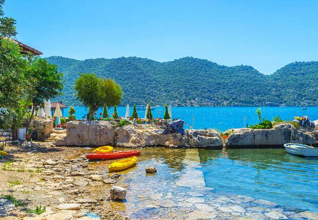 Kayaks rest on a rocky shore beside calm, blue water. Nearby, sun umbrellas and trees provide shade, while green hills rise in the background under a clear blue sky.