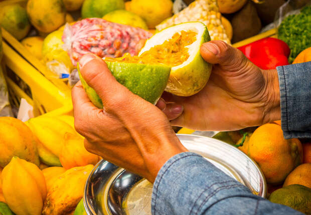 Hands holding a halved passion fruit, exposing juicy seeds, in a market environment. Bright fruits and vegetables surround, with a stainless steel bowl underneath, capturing the fruit’s contents.