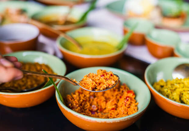 A hand holds a spoon filled with reddish-orange paste over a green-rimmed bowl, surrounded by various colorful dishes in bowls, suggesting a meal preparation or buffet setting.