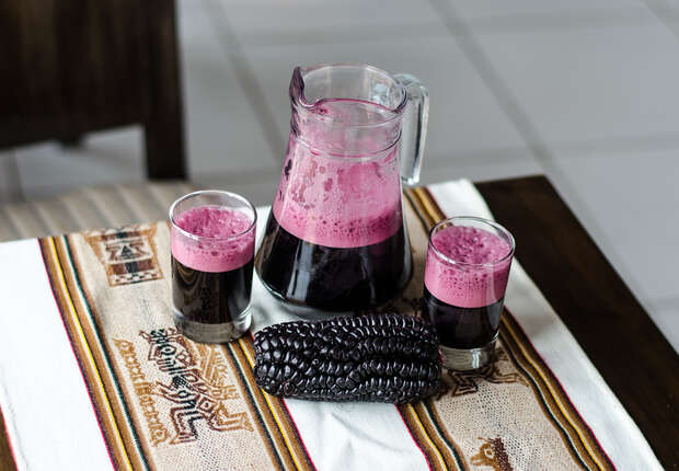 A pitcher and two glasses hold frothy, dark purple liquid, likely juice. The setup includes a cob of purple corn on a colorful woven table runner in a room setting.