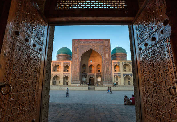 Ornate wooden doors frame a grand building with turquoise domes and intricate tilework. People walk and sit within a spacious, sunlit courtyard, creating a vivid architectural scene.