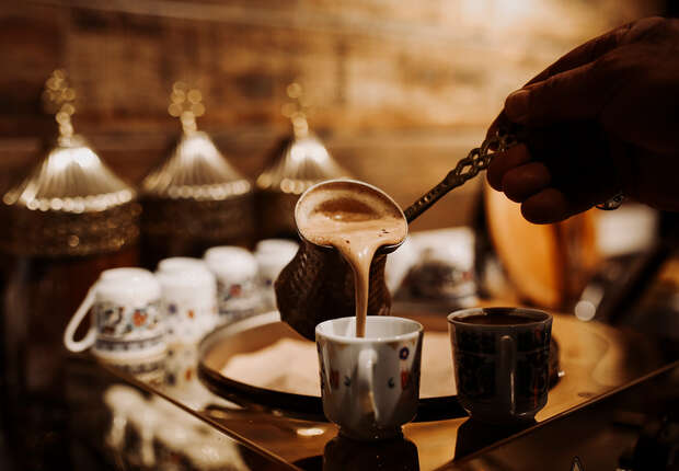 A hand pours coffee from a cezve into a small cup, set on a reflective surface. The background features decorative cups and ornate containers, creating a warm, traditional ambiance.