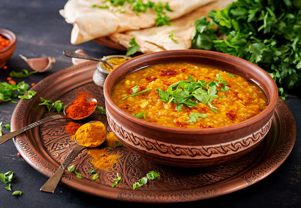 A bowl of lentil soup garnished with cilantro sits on a decorative platter with spoons of spices; flatbread and fresh cilantro are arranged nearby.