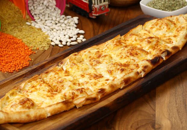 A long, oval-shaped cheese pide rests on a wooden board. In the background, lentils, beans, and grains are scattered beside a toy train on a wooden surface.