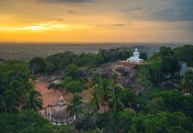 White Buddha statue sits atop a rocky hill surrounded by lush green trees and a temple structure below, under a golden sunset sky extending into the horizon.