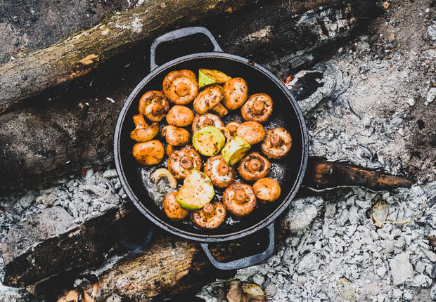 Mushrooms and lemon wedges sizzle in a black pan over a campfire, surrounded by ash-covered logs in an outdoor setting.
