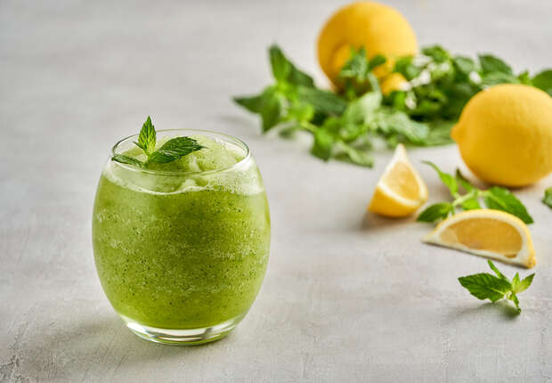 A green smoothie garnished with mint leaves sits in a glass on a gray surface. In the background, whole lemons, lemon wedges, and fresh mint leaves are scattered.