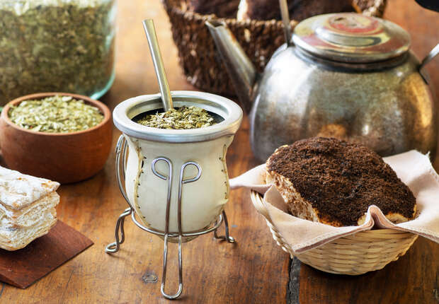 A yerba mate gourd with a straw rests on a wooden table, surrounded by loose leaf herbs, a metal teapot, and bread in a basket.