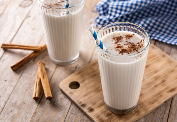Two glasses of creamy, spiced milk with cinnamon on top have striped straws and sit on a wooden board. Nearby are cinnamon sticks and a blue checkered cloth on a wooden table.