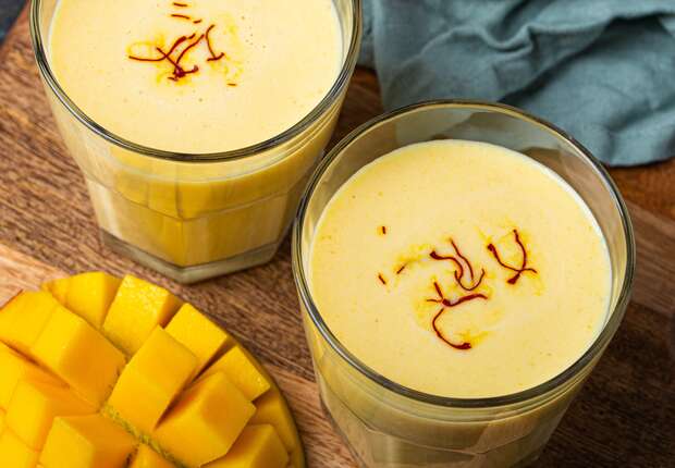 Two glasses of creamy mango lassi topped with saffron strands rest on a wooden surface, alongside diced fresh mango and a teal napkin.