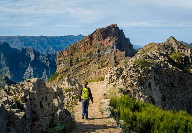 A person with a yellow backpack walks along a rocky mountain path, surrounded by rugged cliffs and distant peaks under a partly cloudy sky.