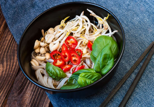 A black bowl holds noodles topped with bean sprouts, sliced red chilies, and fresh basil leaves. The bowl rests on a textured blue cloth, with wooden chopsticks nearby.