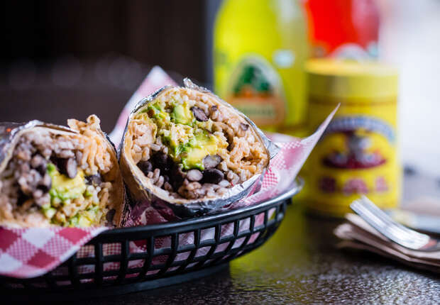 A foil-wrapped burrito, cut in half, reveals rice, beans, and avocado on a red-checkered napkin inside a black basket. Background includes bottled drinks and condiment containers.