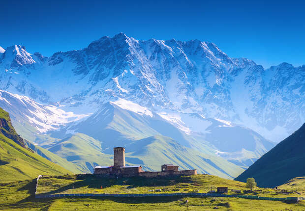 A stone tower stands amidst green grass, surrounded by a vast mountain range with snow-capped peaks under a clear blue sky.