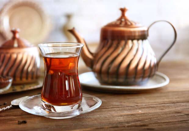 A glass of tea sits on a saucer, with a copper teapot in the background, resting on a wooden table, creating a warm and rustic atmosphere.