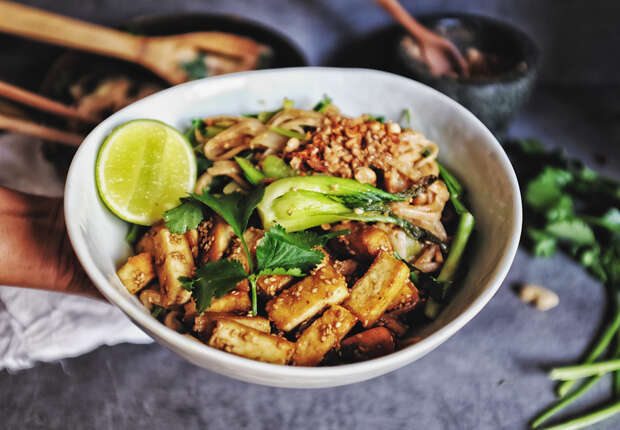 A bowl of stir-fried noodles topped with tofu, greens, peanuts, and a lime wedge is held in one hand, set against a gray surface with blurred kitchen items in the background.