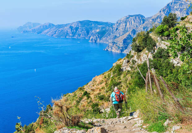 A hiker walks downhill with trekking poles along a coastal trail, surrounded by rugged cliffs and lush vegetation, overlooking a bright blue sea under a clear sky.