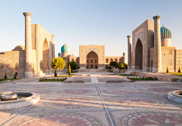 Ancient architectural complex with tall, ornate buildings and blue domes stands under a clear sky. Patterned tiles cover the spacious courtyard, framed by trees and pathways, evoking a historical setting.