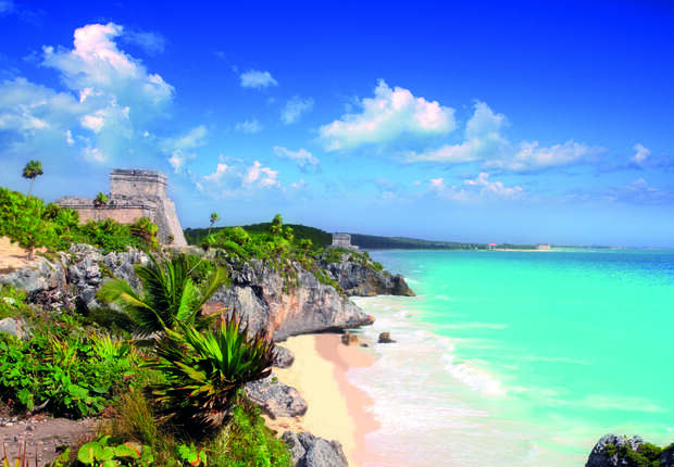 Ancient stone ruins stand atop a rocky cliff, overlooking a turquoise ocean and sandy beach, surrounded by lush green vegetation under a clear blue sky with scattered clouds.