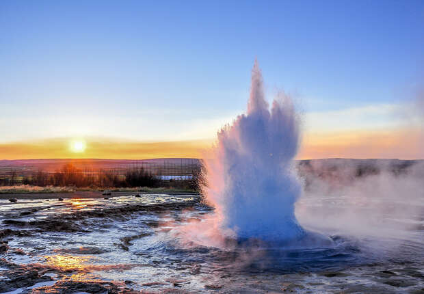 A geyser erupts, spewing water and steam upwards, surrounded by a rocky terrain. In the background, the sun sets on the horizon, casting a warm, golden light.