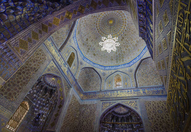 Ornate ceiling displays intricate geometric patterns and blue accents, with a central chandelier. Surrounding walls feature elaborate tilework and decorative arches, suggesting a grand, historic interior space.