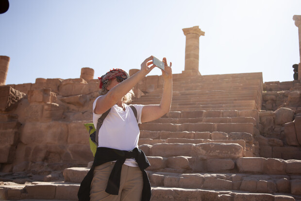 Person taking a selfie, holding a smartphone upward, while standing on ancient stone steps in a historical site with pillars and ruins under a bright, clear sky.