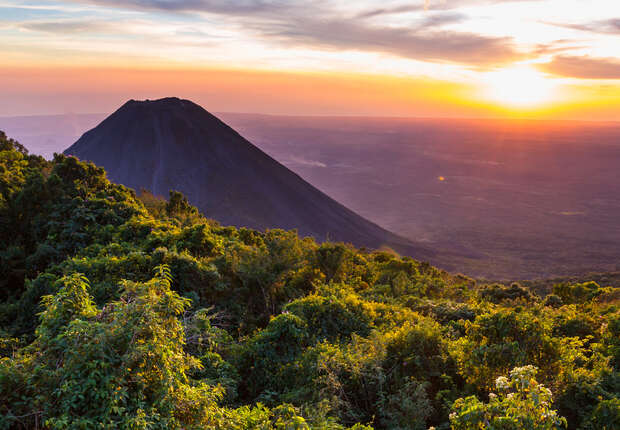 A conical mountain stands under a vibrant sunset, surrounded by lush, green forests. The sky features orange and purple hues, adding warmth to the expansive landscape below.