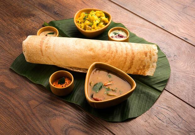 A large, crispy dosa rests on a banana leaf, accompanied by small bowls of chutneys and sambar on a wooden table, creating a traditional South Indian meal setting.