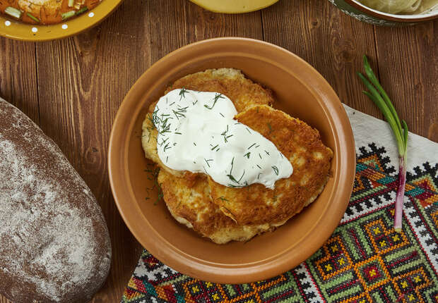 Fried potato pancakes topped with sour cream and dill sit on a brown plate. Nearby, a dusted loaf of bread rests on a colorful patterned cloth over a wooden table.