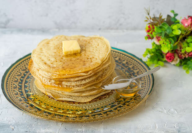 A stack of pancakes topped with butter and drizzled with syrup sits on an ornate plate. A small jar of honey with a spoon is nearby, and a bouquet of flowers decorates the background.