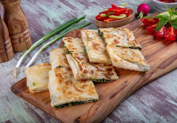 Flatbreads filled with greens are stacked on a wooden board. Near them, sliced peppers and fresh vegetables add vibrant color, with wooden salt and pepper mills in the background.