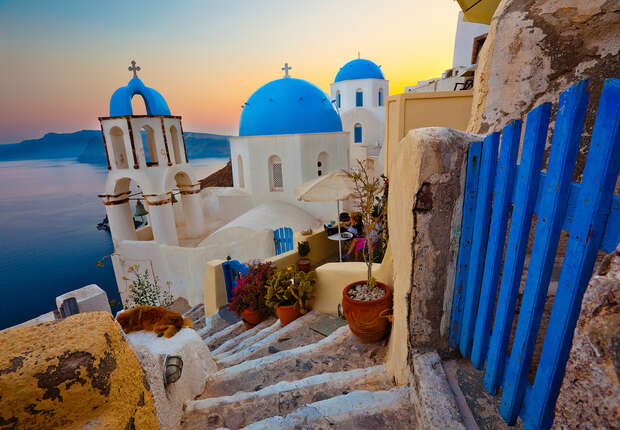 White buildings with blue domes sit beside a stone staircase adorned with potted plants. A calm sea and warm sunset hues create a serene coastal backdrop. A blue gate outlines the path.