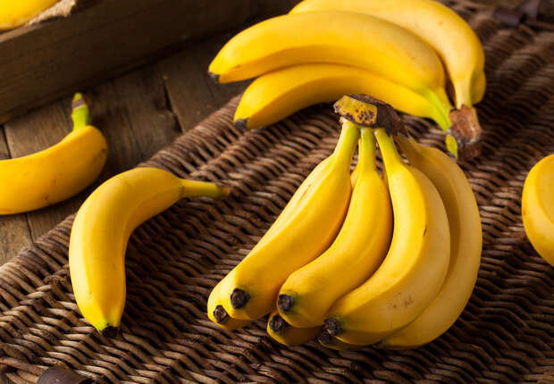 Ripe bananas rest on a woven surface, some in a bunch and others scattered. In the background, a wooden box and more bananas are visible, creating a rustic setting.