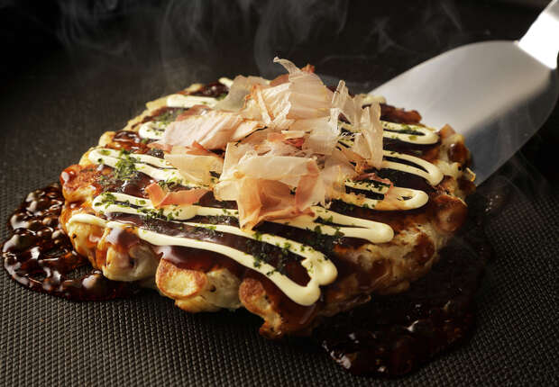 A savory pancake topped with mayonnaise, sauce, and bonito flakes, sits steaming on a hot grill. A metal spatula is placed nearby, ready for serving.