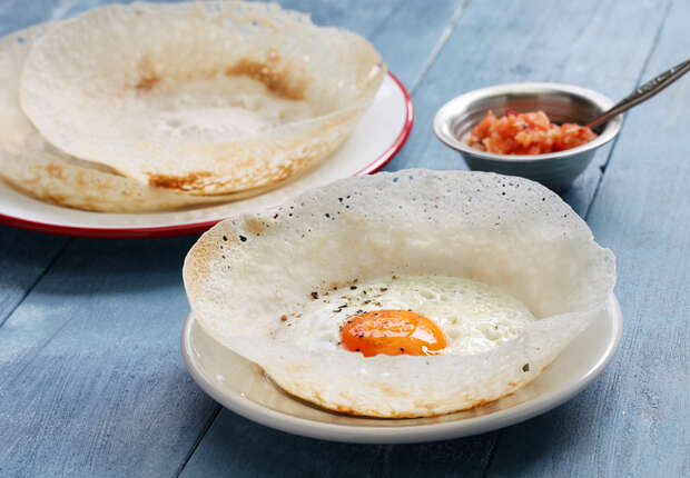 A crispy crepe with a sunny-side-up egg rests in a shallow white dish, accompanied by tomato chutney in a small bowl on a blue wooden table.