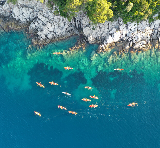 Kayaks float in clear turquoise water near a rocky coastline with lush greenery, creating a vibrant contrast between the deep blue sea and the rugged, verdant shoreline.