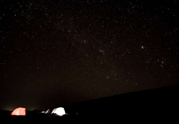 Tents softly illuminated, sit quietly under a vast, star-filled night sky, offering a serene camping experience in an open natural setting.