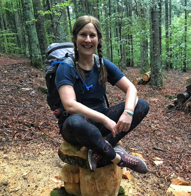 A person sits on a carved wooden stump, smiling, wearing outdoor clothing and backpack, in a forest with trees and fallen leaves, suggesting a hiking activity.